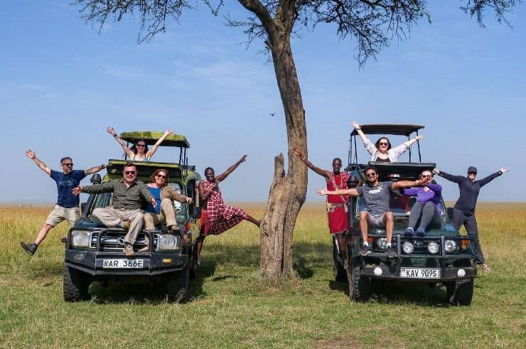 Happy safari group celebrating with Maasai guide beside 4×4 Land Cruisers in the Masai Mara, Kenya